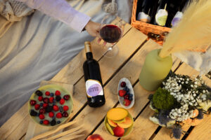 Eine Flasche alkoholfreier Rotwein Zeronimo Leonis Blend auf einem Palettentisch daneben ein Glas mit Rotwein und eine Hand, die zu diesem Glas greift, Blumen und Früchte liegen auch auf der Palette